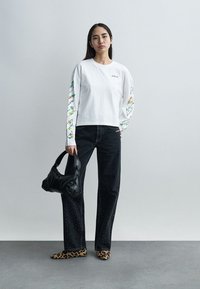 Γυναίκα που φοράει λευκό μπλουζάκι μακρυμάνικο με φλοράλ μανίκια, μαύρα τζιν με τρουκς, παπούτσια με λεοπάρ σχέδιο, κρατώντας μια μαύρη τσάντα.