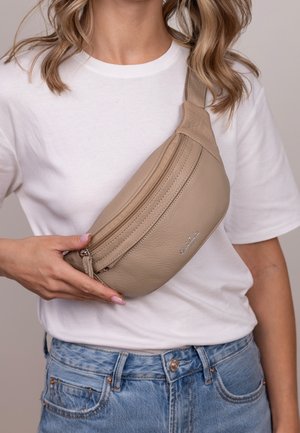 Mujer con camiseta blanca y vaqueros azules que lleva un bolso cruzado de cuero beige con cremallera sobre el pecho.