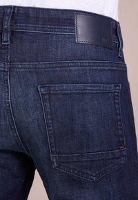 Tmavě modré džíny z denimu s koženou záplatou na pasu, několika stehy a jednou zadní kapsou s jemně zakřiveným tvarem.