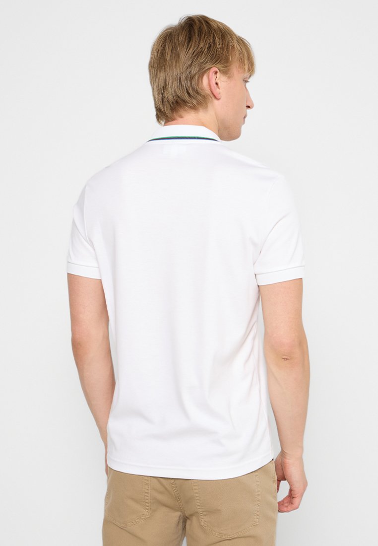 Homme blond portant un polo blanc à manches courtes uni et un pantalon beige, de dos contre un fond clair.