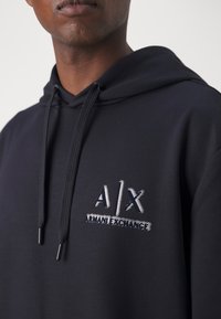 Mörkblå hoodie med en framficka i känguru-stil, prydd med en texturerad A|X-logga i vitt och silverbroderi på bröstet. Mjuk tygkänsla och justerbara snoddar.