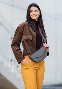 Šedá kožená crossbody taška s prohnutým klopou, zkombinovaná s hnědou kordovou bundou a hořčicově žlutými kalhotami, které ukazují moderní outfit.