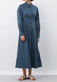 Abito lungo, con bottoni, di un profondo colore teal. Realizzato in un tessuto morbido, presenta un colletto, plissettature e maniche lunghe. Indossato con sandali leggeri.