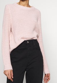 Helles rosa gestricktes Sweatshirt mit Rundhalsausschnitt, strukturiertem Muster und lockerer Passform, kombiniert mit hochgeschnittenen schwarzen Jeans.