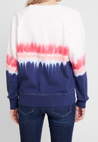 Personne portant un sweat-shirt à bandes tie-dye horizontales en blanc, rouge, rose et bleu marine, associé à un jean denim bleu foncé.
