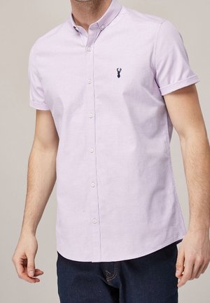 Camisa de manga corta de color púrpura claro con cuello abotonado; presenta un pequeño logo en navy en el pecho y un dobladillo recto. El material parece suave.