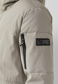 Beige gesteppte Jacke mit einem Reißverschluss-Ärmeltasche und einem schwarzen Etikett mit weißem Text. Glattes, strukturiertes Gewebe mit dezentem Glanz.