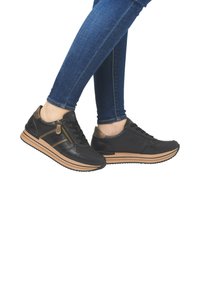 Schwarze Leder-Sneaker mit einem braunen Akzent, ausgestattet mit einem seitlichen Reißverschluss, strukturierten Einsätzen und einer gerippten Sohle. Getragen mit blauen Jeans.