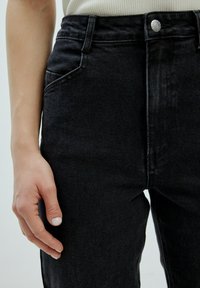 Schwarze Jeans aus Denim mit hoher Taille, ausgestattet mit einem Knopfverschluss, Fronttaschen und sichtbaren Nähten. Glatte Textur und eng anliegende Passform.
