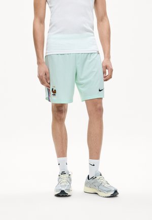 Άνδρας που φοράει ανοικτό πράσινο ποδοσφαιρικό σορτς της Γαλλίας με το έμβλημα FFF και το λογότυπο της Nike, λευκές κάλτσες Nike και γκρι αθλητικά παπούτσια Nike.