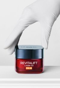 Crème Revitalift Laser X3 dans un pot en verre. Base rouge avec un couvercle bleu. L'étiquette affiche 'FPS 25' en orange, tenue par une main gantée.