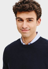 Ciemnoniebieski sweter o teksturowanym splocie, noszony na jasnoniebieskiej koszuli z kołnierzykiem. Model ma kręcone włosy i się uśmiecha.