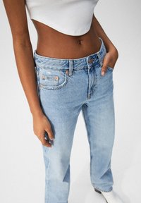 Jeans de mezclilla azul claro con corte recto. Presentan costuras naranjas, bolsillos frontales y un cierre de botón visible. Textura suave.