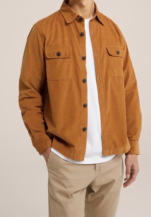 Homme portant une chemise en velours côtelé marron boutonnée sur un t-shirt blanc avec un pantalon beige, debout avec une main dans la poche.