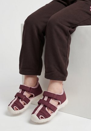 Kind zit op een witte blok, draagt een donker broek en wijnrode open-toe sandalen met twee verstelbare banden en witte zolen.