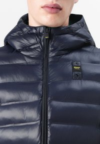 Navy puffer jacket with a high collar, horizontal quilted design, front zipper, and branded tag on the upper left side. Smooth texture.