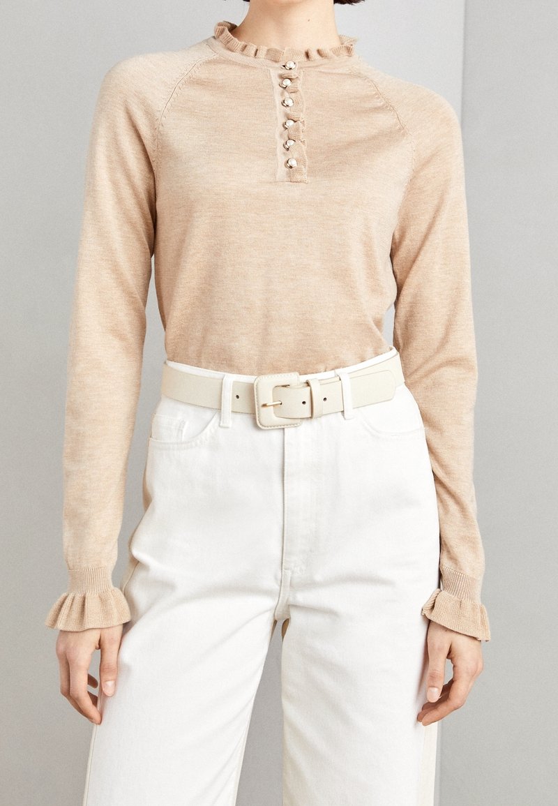 Femme portant un top beige à manches longues avec des poignets à volants et des boutons en perles, associé à un pantalon blanc taille haute et une ceinture blanche.