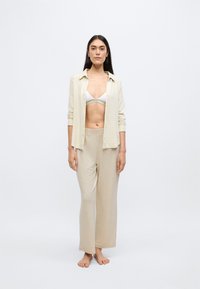 Bézs kötött ing gombokkal, fehér melltartó és bézs széles szárú nadrág fölött viselve. A ruházat laza szabású és puha tapintású.