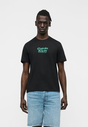 Νέος άνδρας με σκούρο δέρμα και πλεξούδες, φορεί μαύρο t-shirt Calvin Klein και ανοιχτόχρωμα τζιν σορτς, μπροστά σε απλό φόντο.