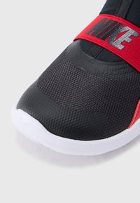 Chaussure de sport noire avec un matériau texturé et une semelle blanche. Elle présente une sangle rouge avec le logo « NIKE » sur le dessus.