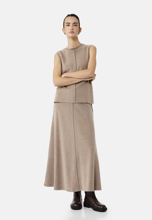 Femme debout, les bras croisés, portant un haut sans manches beige, une jupe longue assortie et des bottines marron foncé.