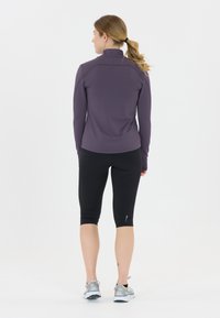 Violetti pitkähihaisen urheiluvaatteen, jossa on korkea kaulus, yhdistetty mustiin capri-legginseihin ja harmaisiin juoksukengiin. Yksinkertainen kangas ilman kuvioita.