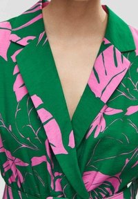 Blusa verde con grandi motivi di foglie tropicali rosa e colletto a V profonda, indossata da una persona che mostra le spalle e la parte superiore del petto.
