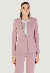 Donna che indossa un completo giacca e pantaloni rosa con una maglietta bianca decorata con dettagli rosa e perle, in piedi di fronte a uno sfondo neutro.