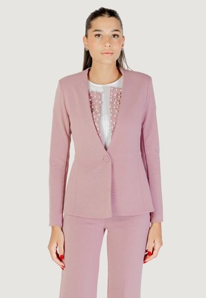 Donna che indossa un completo giacca e pantaloni rosa con una maglietta bianca decorata con dettagli rosa e perle, in piedi di fronte a uno sfondo neutro.