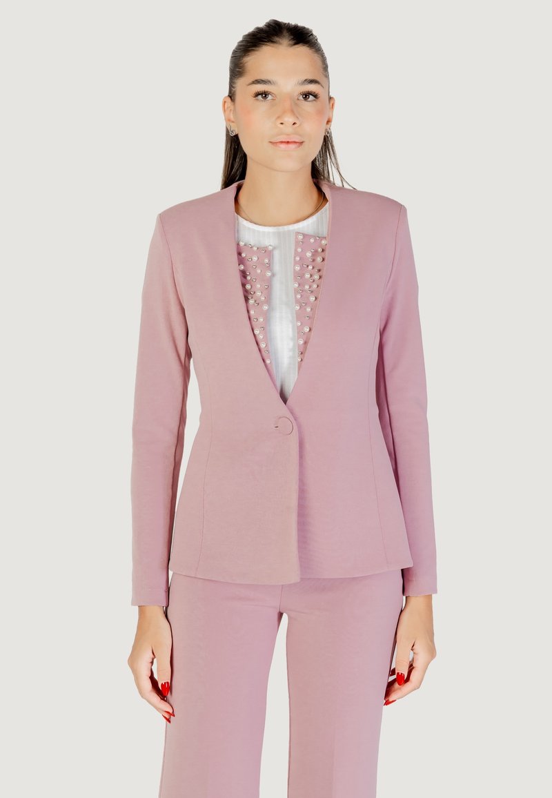 Donna che indossa un completo giacca e pantaloni rosa con una maglietta bianca decorata con dettagli rosa e perle, in piedi di fronte a uno sfondo neutro.