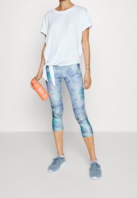 Camisa blanca de manga corta con detalle de nudo; leggings teñidos con efecto tie-dye en azul y verde; zapatos deportivos grises; botella de agua naranja.