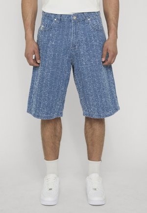 Homme portant un short bleu texturé jusqu'aux genoux, des chaussettes blanches, des baskets blanches et une chemise blanche, debout devant un fond uni.