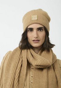 Beige gebreide muts met een geribbelde textuur en een klein decoratief label, gecombineerd met een bijpassende geribbelde sjaal. De stof lijkt zacht en dik.