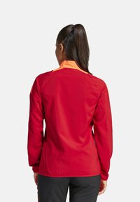 Veste de sport rouge avec un col orange, texture lisse et manches longues ; elle comporte des poches latérales et a une coupe ajustée.