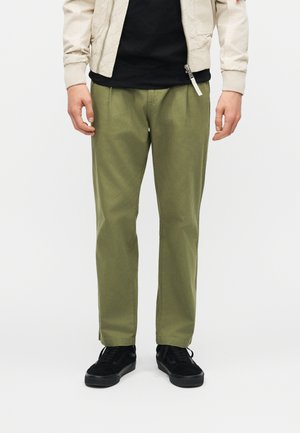 Uomo con pantaloni plissettati verde oliva, sneaker nere, maglietta nera e giacca beige chiaro con zip, in piedi su sfondo bianco.