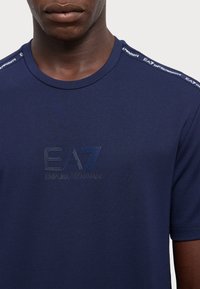 Navy blauwe t-shirt met ronde halslijn, korte mouwen en EA7-logo in ton-sur-ton stiksel aan de voorkant. Accenten op de schouders met branding.