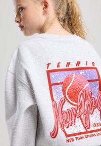 Gray sweatshirt featuring a retro-style print of a tennis ball and "New York Sports Academy" in vibrant red and purple graphics.