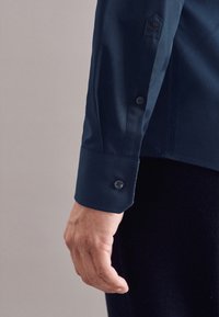 Marineblaue Hemdsärmel mit Knopfmanschetten, versehen mit einer gestickten Verzierung an der Schulter und glatter Stofftextur. Die Hand ist in der Nähe des Ärmels positioniert.