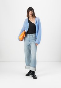 Lichtblauwe fuzzy cardigan over een zwarte top, gecombineerd met opgerolde lichtblauwe jeans en zwarte schoenen. Houdt een oranje handtas vast.