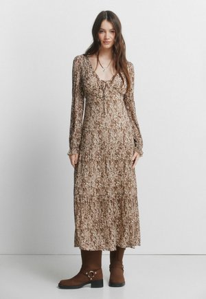 Vrouw die een bruine maxi-jurk met lange mouwen en patroon draagt, met een strikdetail en bruine laarzen tot halverwege de kuit, staand tegen een effen grijze muur.