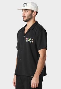 Svart kortermet skjorte med krage og fargerik "PASSE DE MONACO" tekst med blomsterdetaljer brodert på venstre bryst.