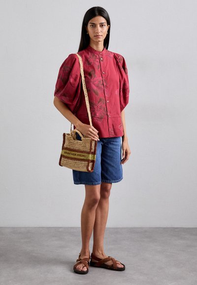 Blouse rouge à motifs floraux avec des manches bouffantes, associée à un short en denim et un sac en osier. Des sandales marron complètent la tenue.