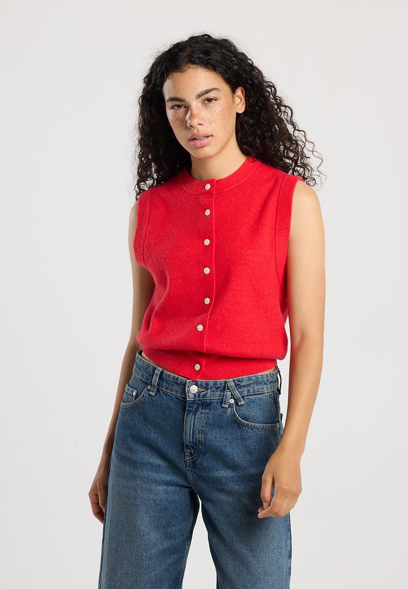Red sleeveless sweater in a soft knit with a round neck and front button closure, paired with blue high-waisted jeans.