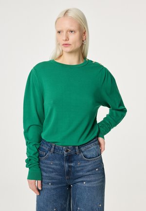 Vrouw met lang blond haar, gekleed in een groene trui met lange mouwen en gouden knopen op de schouder, en blauwe jeans met kleine hartjespatronen.