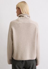 Beżowy sweter dziany z wysokim kołnierzem, o luźnym kroju i prążkowanym wykończeniu. Sweter ma miękką teksturę i detale szwów na ramionach.