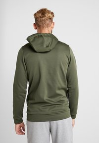 Sudadera de un verde oscuro con un diseño ajustado, capucha ajustable y textura suave, combinada con pantalones de chándal grises, vista desde la parte de atrás.
