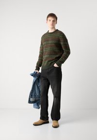 Green and brown patterned sweater, black jeans, tan shoes. Model holds a denim jacket, standing in a neutral space with plain background.