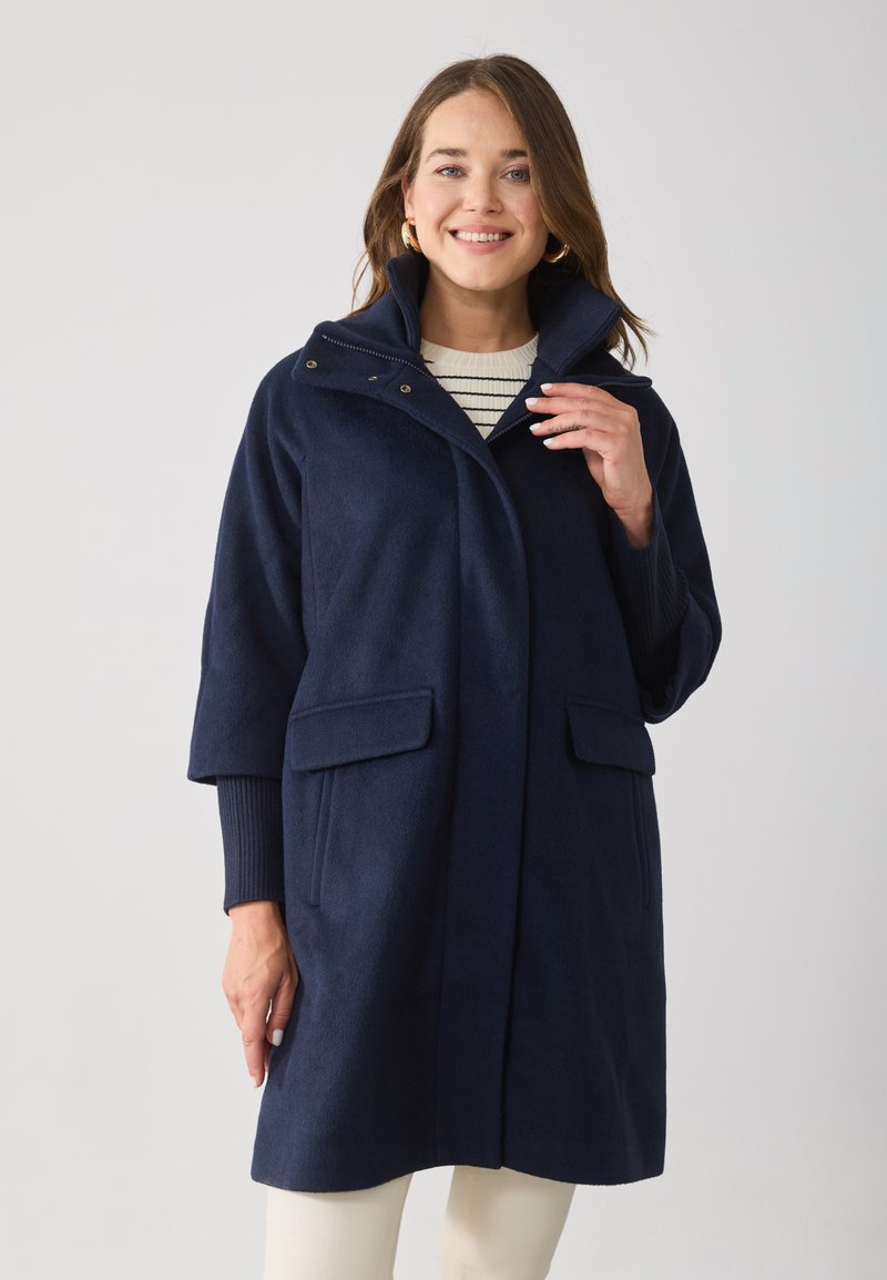 Marineblauer Mantel aus weichem Stoff, mit hohem Kragen, zwei Fronttaschen und lässiger Passform. Mit Druckknopfverschluss akzentuiert.