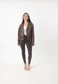 Kobieta stojąca na białym tle, ubrana w oversize'ową brązową skórzaną kurtkę, biały krótki top, czarne obcisłe dżinsy i beżowe sandały na obcasie.