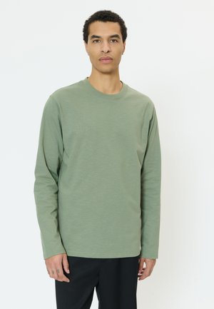 Vīrietis, tērpies gaiši zaļā ar garām piedurknēm kreklā un melnās biksēs, stāv pret vienkāršu baltu fonu, skatās taisni ar neitrālu izteiksmi.